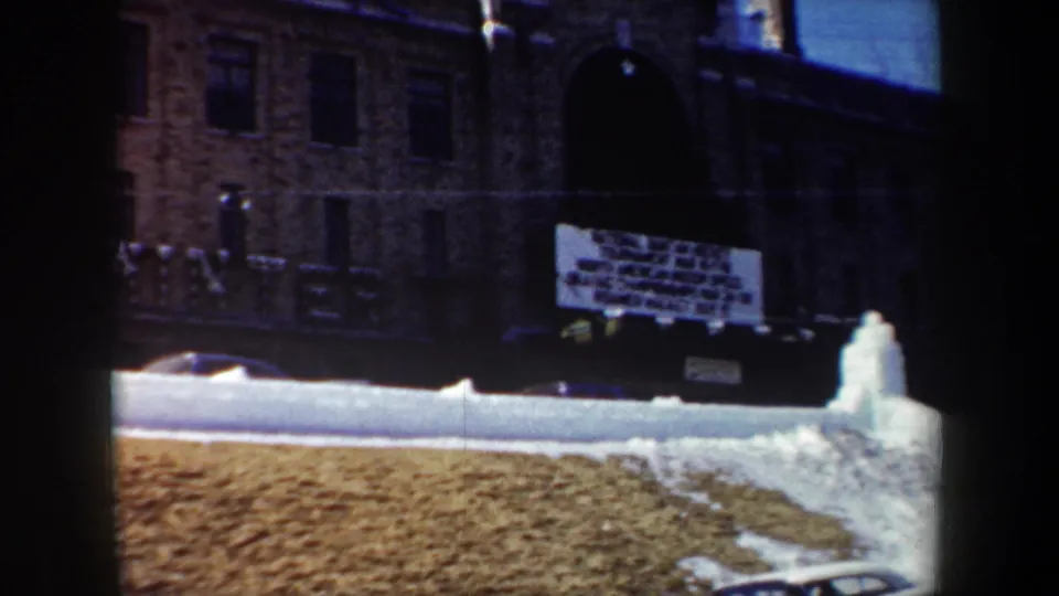 1962 New York home-movie frame of a brick building entrance, white sign, parked car, facade lettering, and piled snow in the foreground.
