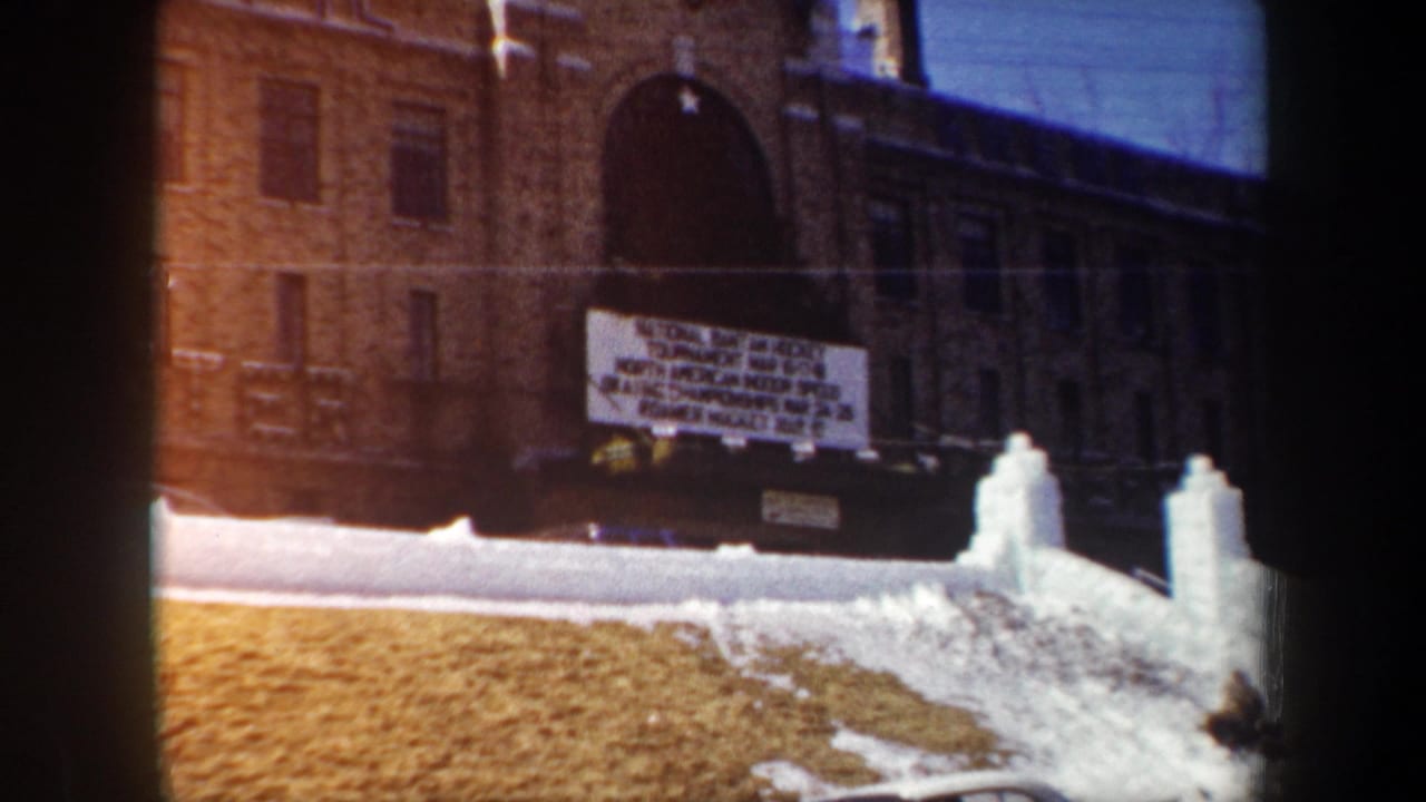 1962 New York 8mm frame showing a brick school exterior, white winter sports tournament sign, and snow-covered entry area.