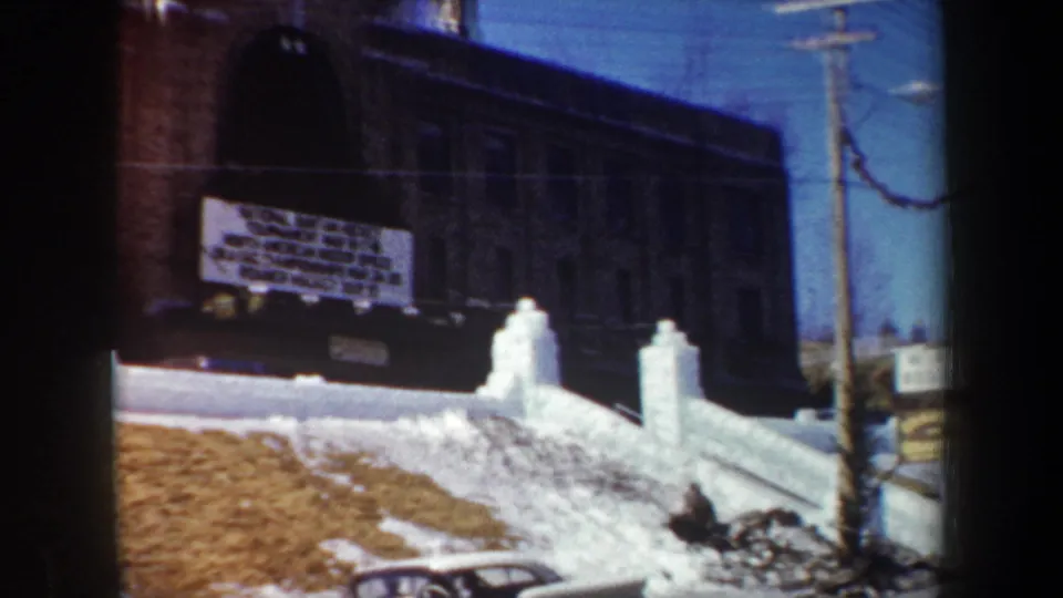1962 New York home-movie frame with a brick school, readable event banner, snowbanks, and roadside utility pole.