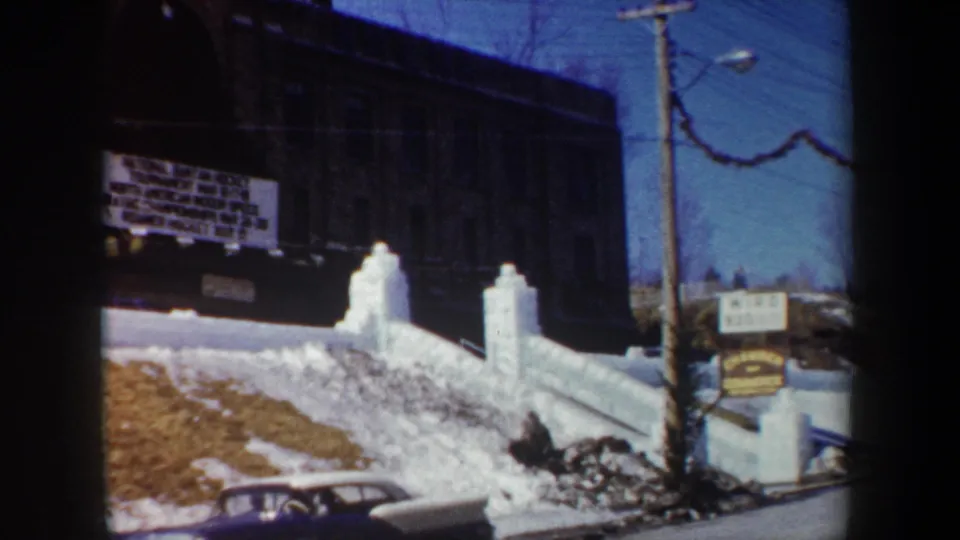 1962 New York 8mm frame showing the school exterior, winter sports sign, snowbanks, roadside sign, and a parked car.