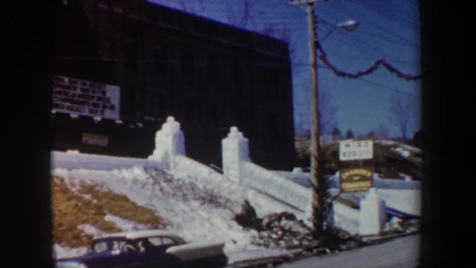 1962 New York home-movie frame with gate shadow, snowy school grounds, event signage, utility pole, and parked car.