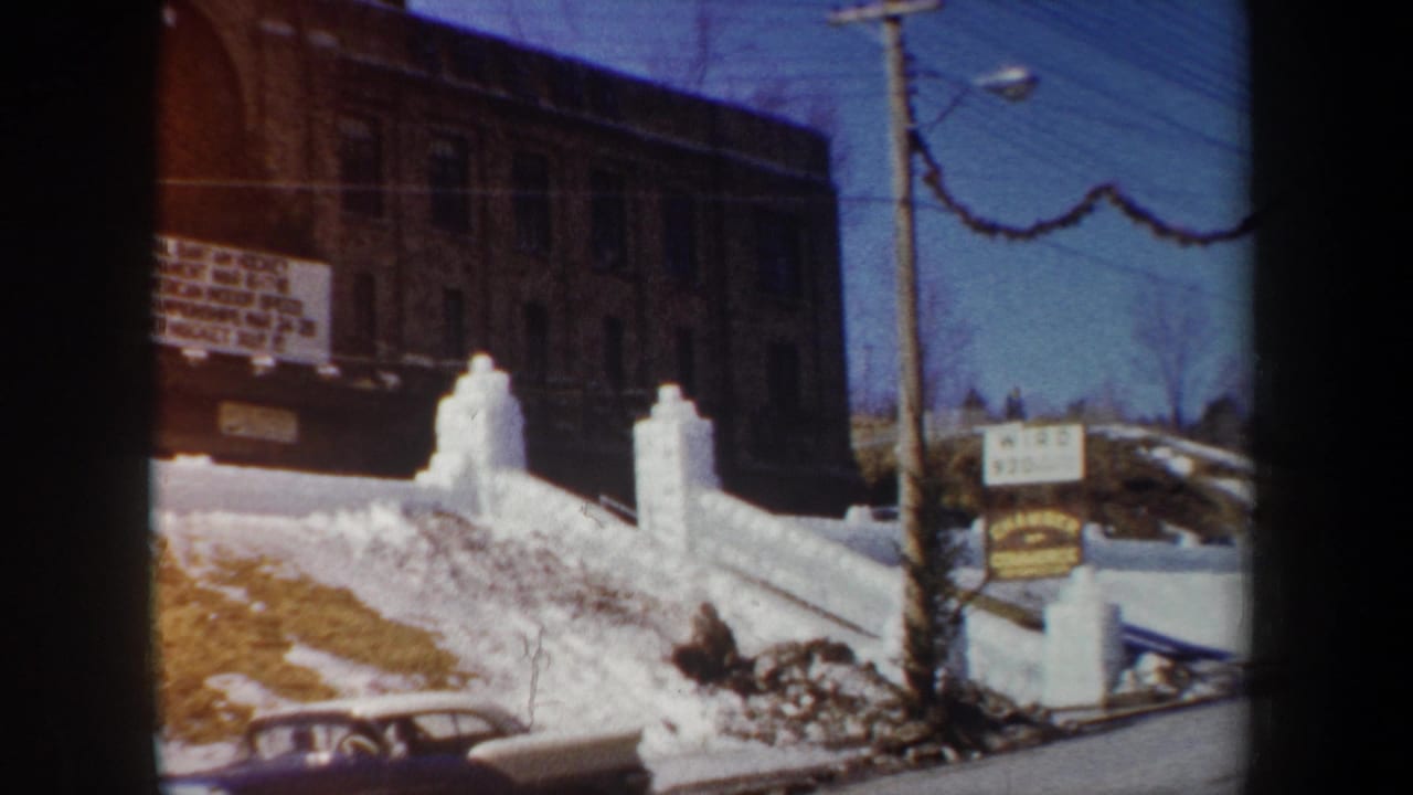1962 New York 8mm frame showing snowbanks, a brick building, a parked car, utility pole, and partially readable event signage.