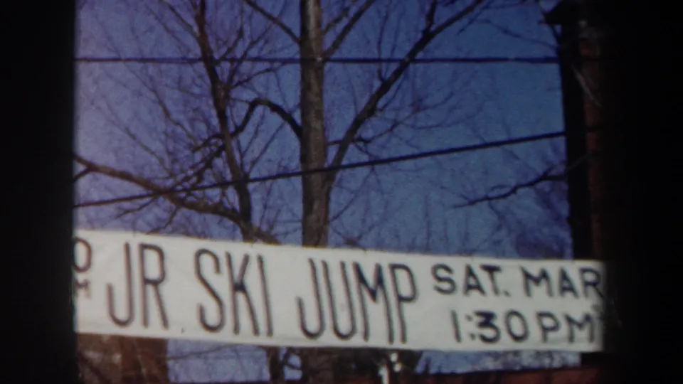Close view of a white JR SKI JUMP banner in a snowy New York winter setting, with bare trees and utility wires behind it.
