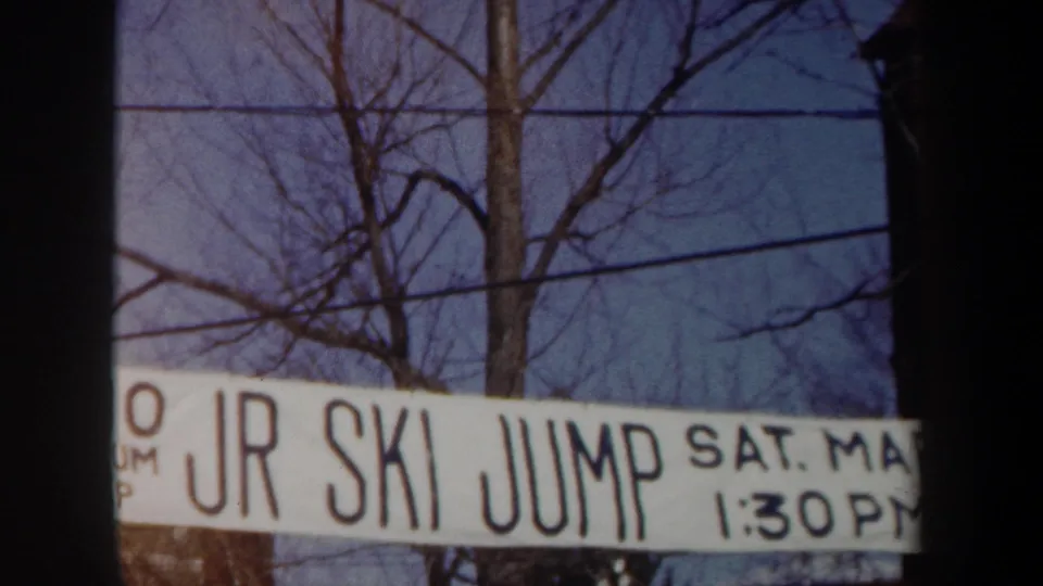 1962 New York frame with the JR SKI JUMP banner partly cropped across the lower view, backed by winter trees and utility wires.