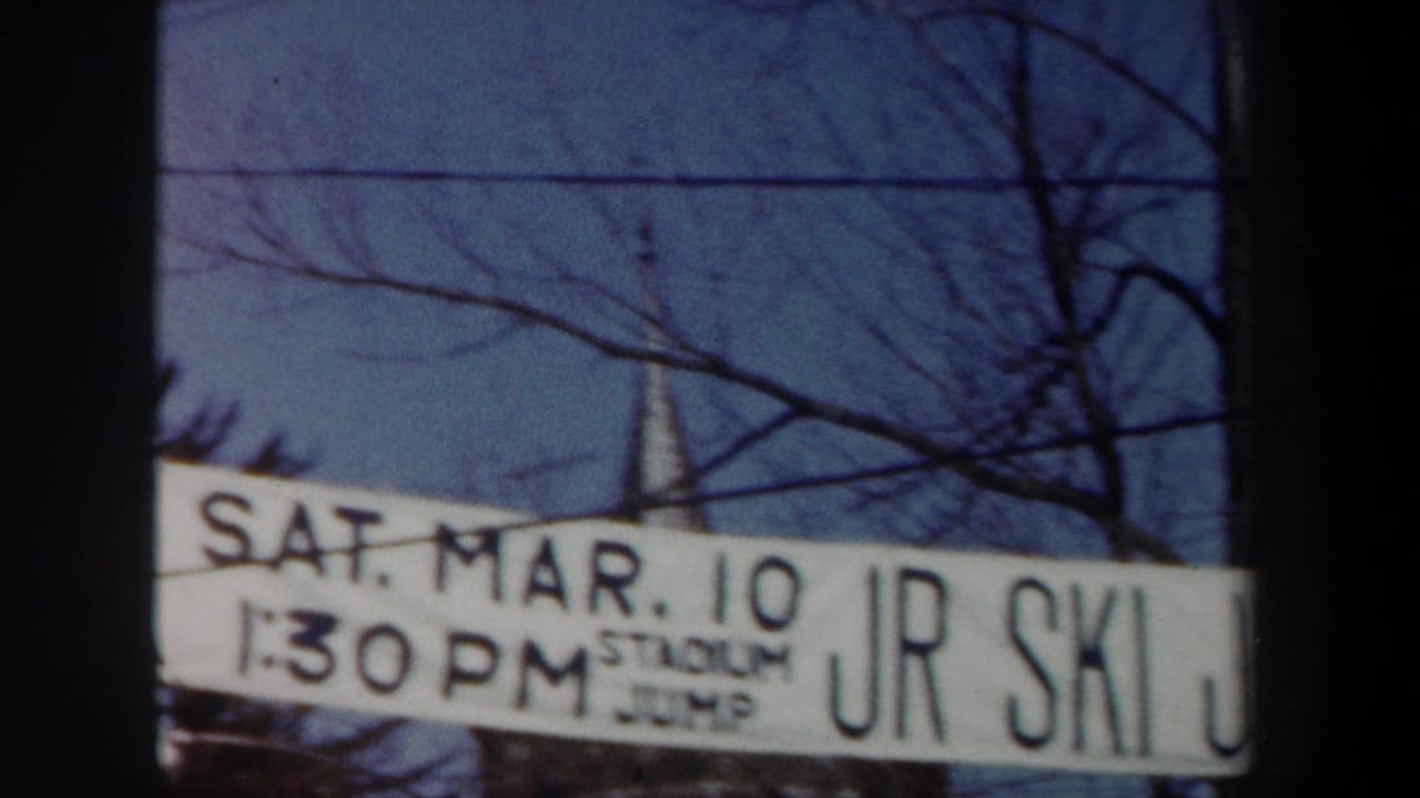 White junior ski event banner above a snowy slope with bare trees and a church steeple behind it.