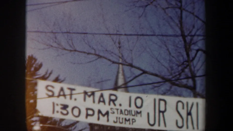 1962 New York home-movie frame with a white banner reading SAT. MAR. 10 and STADIUM JUMP JR SKI near winter trees.