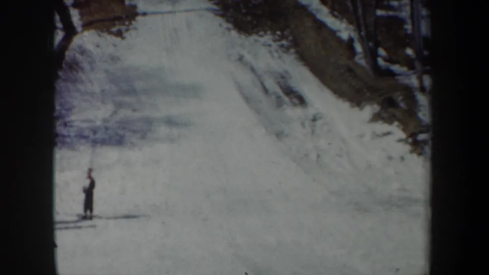 1962 New York 8mm frame showing a snowy ski slope, a distant skier, dark tree trunks, and winter ground cover.