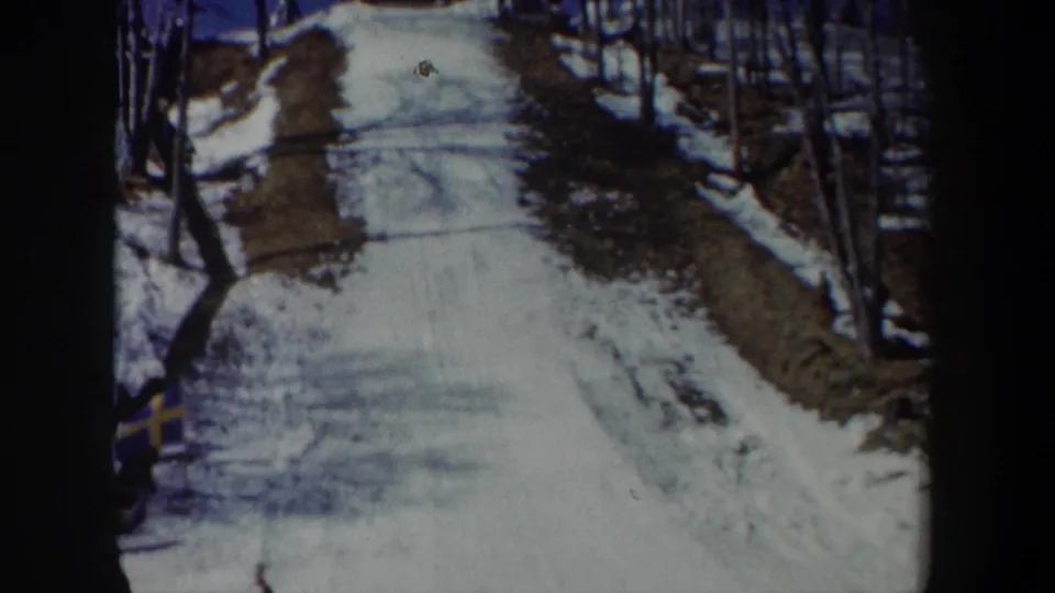 1962 New York home-movie frame looking up a narrow snowy slope bordered by bare winter trees and a distant skier.