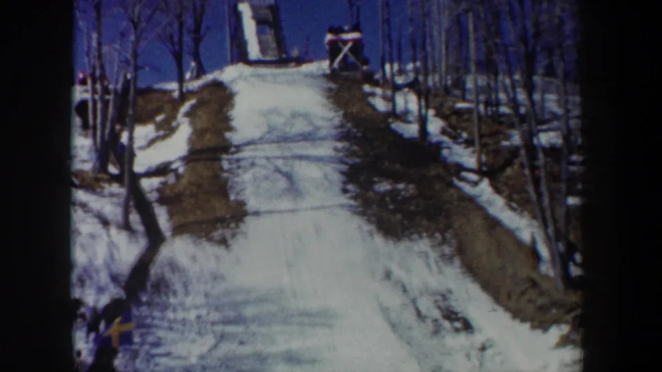1962 New York 8mm frame showing a snow-covered ski-jump slope, bare trees, wooden jump structure, and small Swedish flag.