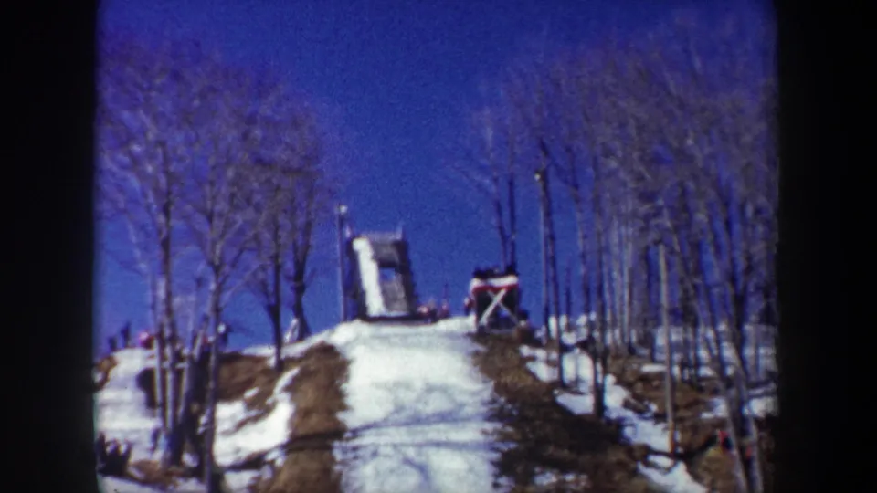 1962 New York 8mm frame looking uphill at a snow-covered ski-jump slope bordered by bare winter trees.
