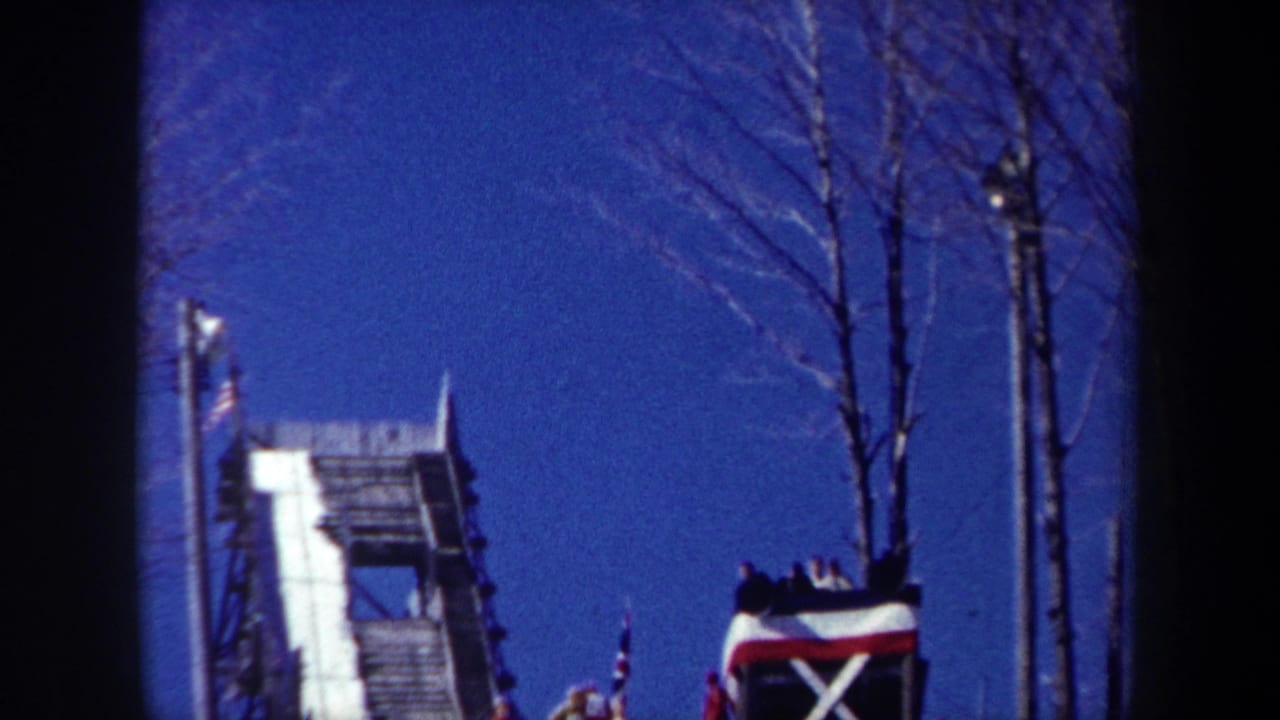 1962 New York home-movie frame showing a snow-lined wooden ramp, bare trees, poles, and spectators clustered near the crest.