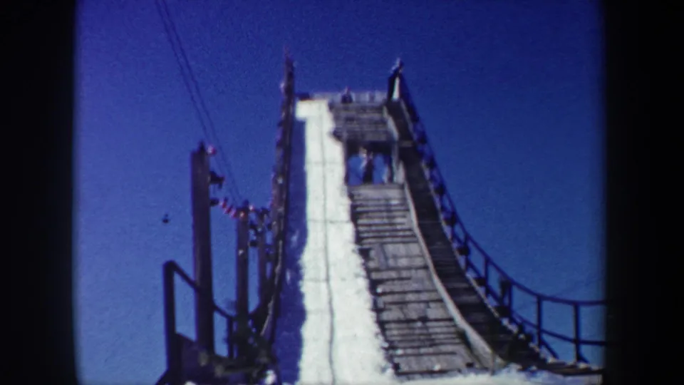 Home-movie view of a steep snow-covered ramp in 1962 New York, with railings, support posts, and people near the upper platform.