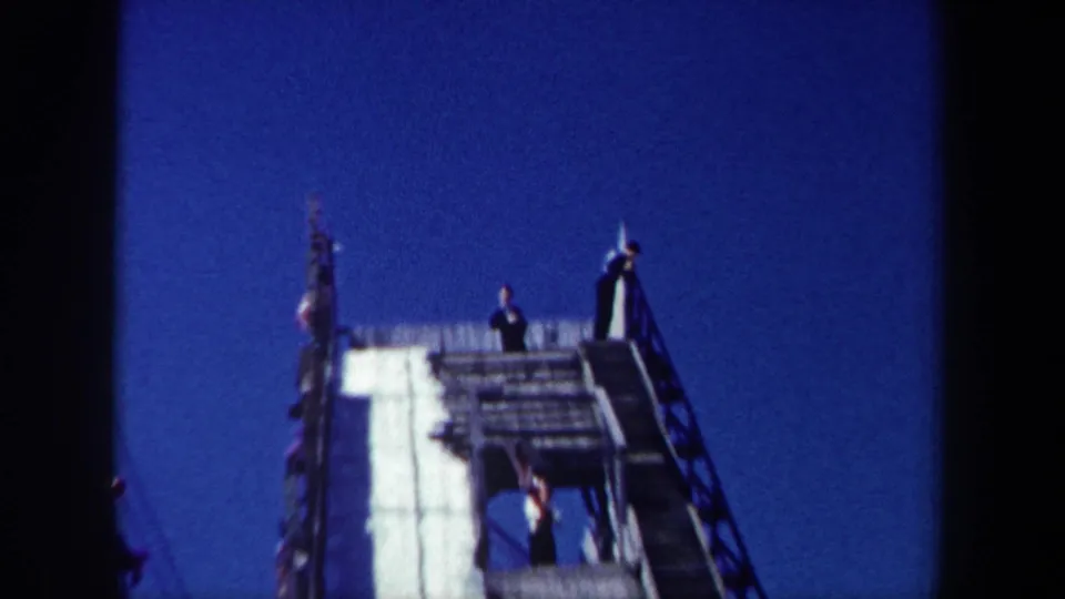 1962 New York frame showing people at the top of a snow-covered wooden ramp, with railings silhouetted against a deep blue sky.