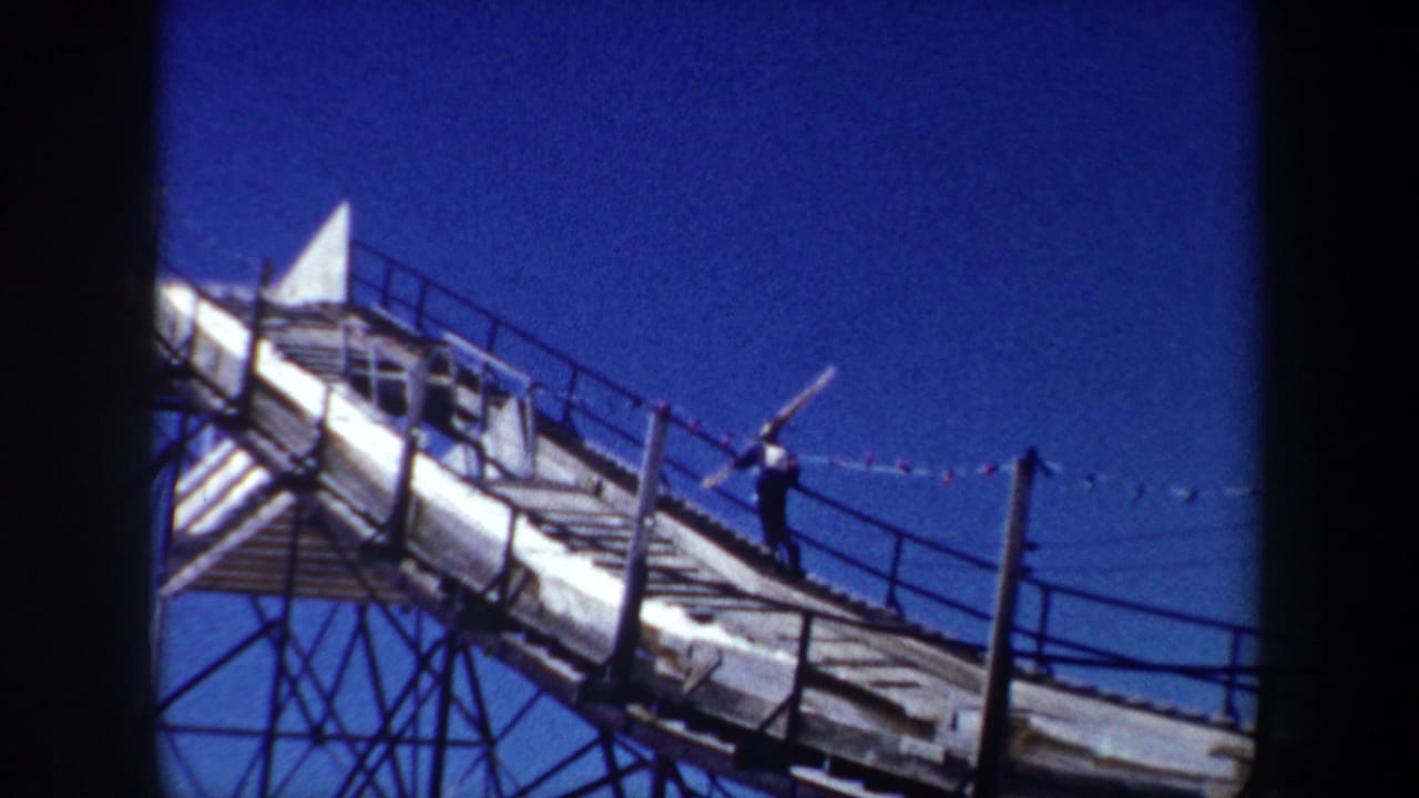 Skier carrying skis up a steep wooden ski-jump ramp beneath a blue sky.
