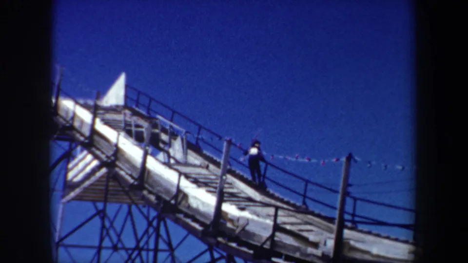 1962 New York home-movie frame of a skier on the upper portion of a wooden ski-jump ramp, framed against clear blue sky.