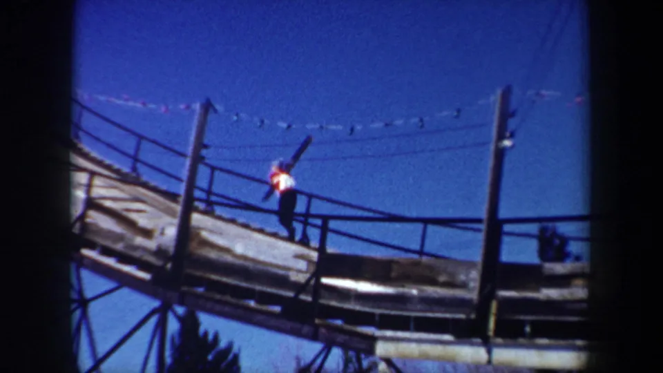 1962 New York home-movie frame of a skier on the ski-jump ramp, holding skis upright near railings and support posts.