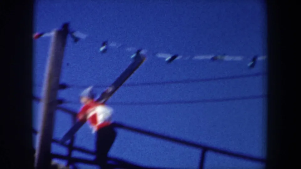 1962 New York frame of a skier moving up resort stairs, with skis, a utility pole, wires, and blue sky visible.