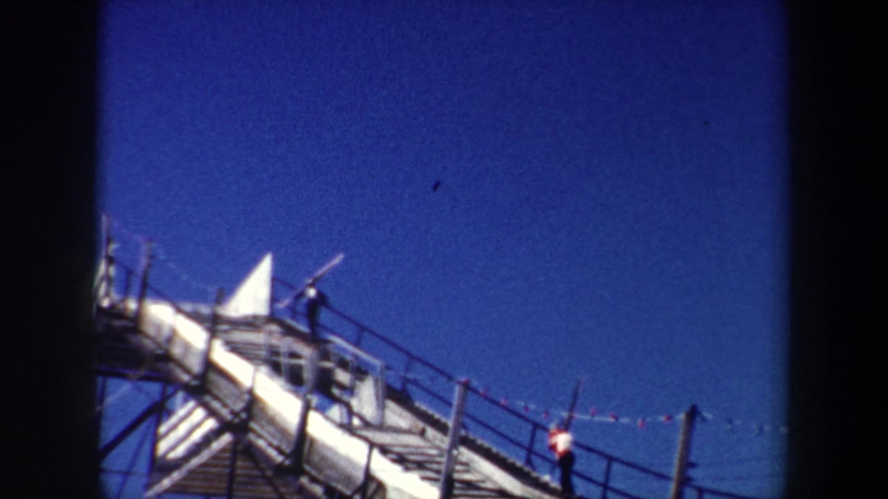 1962 New York 8mm frame showing two men moving along a steep metal walkway with long poles, railings, and blue sky visible.