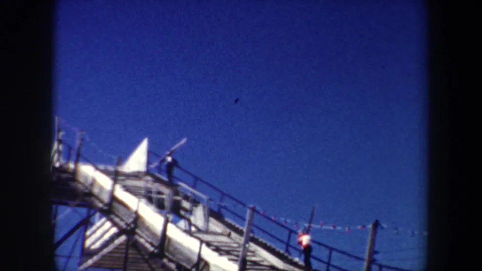1962 New York 8mm frame showing two men moving along a steep metal walkway with long poles, railings, and blue sky visible.
