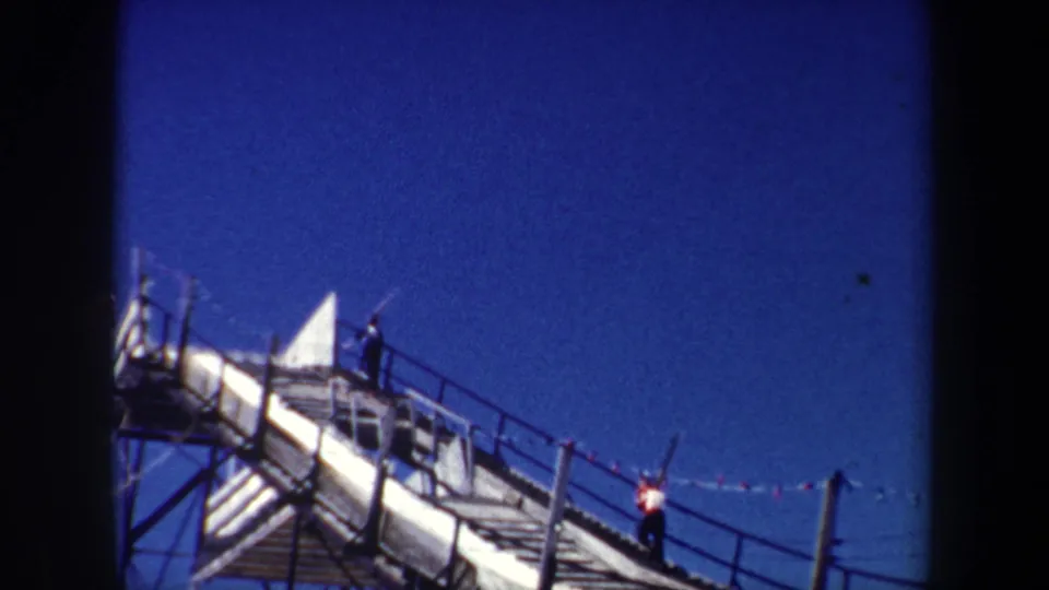1962 New York 8mm frame with workers on a narrow elevated walkway, metal railings, support framing, and open blue sky.