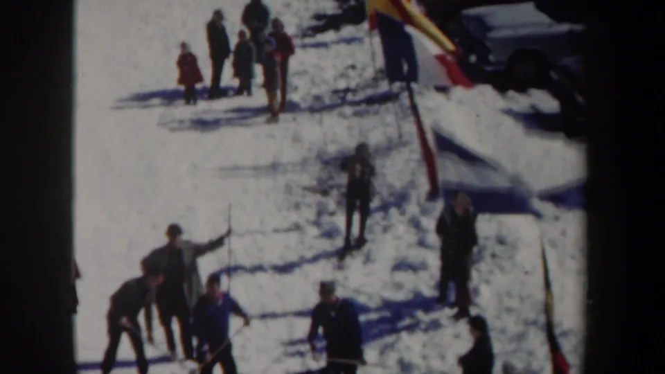 1962 New York home-movie frame with people on snow, marker flags planted nearby, and a partial dark vehicle at the edge.