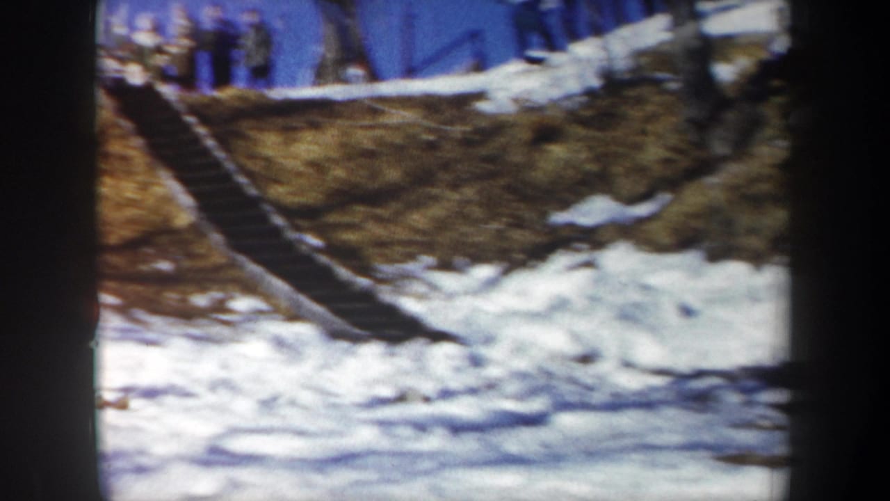 Snowy hillside with a wooden ski-jump ramp and spectators standing along the ridge.