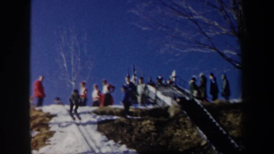1962 New York home-movie frame showing bundled spectators near a wooden ski-jump ramp under a clear blue sky.
