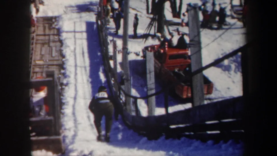 1962 New York ski-area frame with a dark-clothed skier on a snowy path, trees, spectators, and red resort equipment nearby.