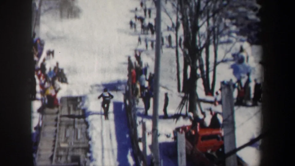 1962 New York ski-resort frame showing a skier raising both arms on the snow with spectators, trees, and red equipment around the slope.