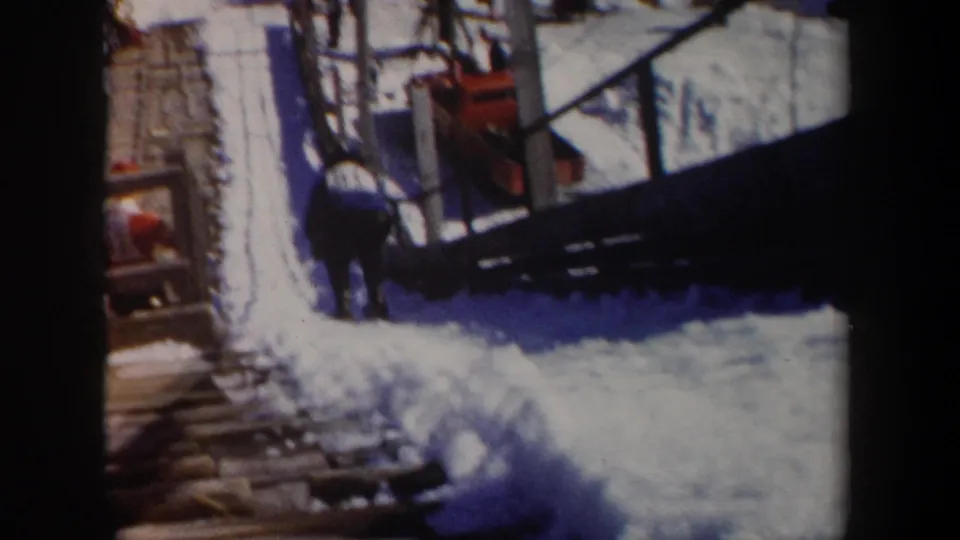 1962 New York home-movie frame showing a snowy ski-area approach with wooden steps, railings and a bundled person near the structure.