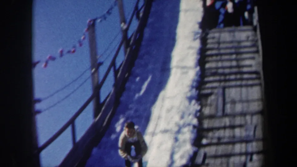 1962 New York ski-jump setting with a snowy ramp, side railings, small flags, wooden steps and a person at the bottom of frame.