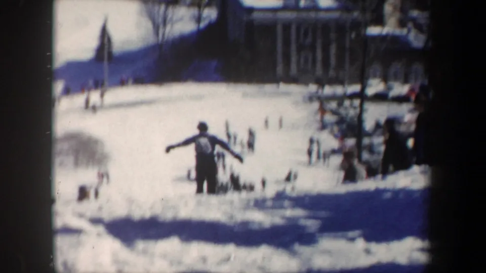 1962 New York ski competition frame showing a skier with arms extended on a bright snowy slope, with distant spectators and buildings.