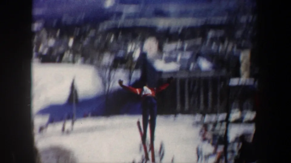 Bibbed skier in red and blue appears airborne or mid-glide on a snowy New York slope, captured in soft 8mm color in 1962.
