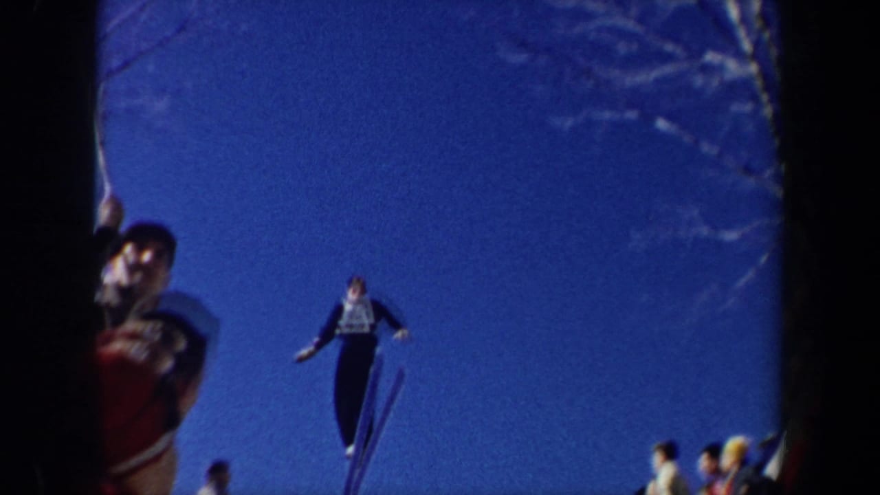 1962 New York 8mm frame showing a skier in a dark outfit and numbered bib mid-jump against a deep blue sky with spectators below.
