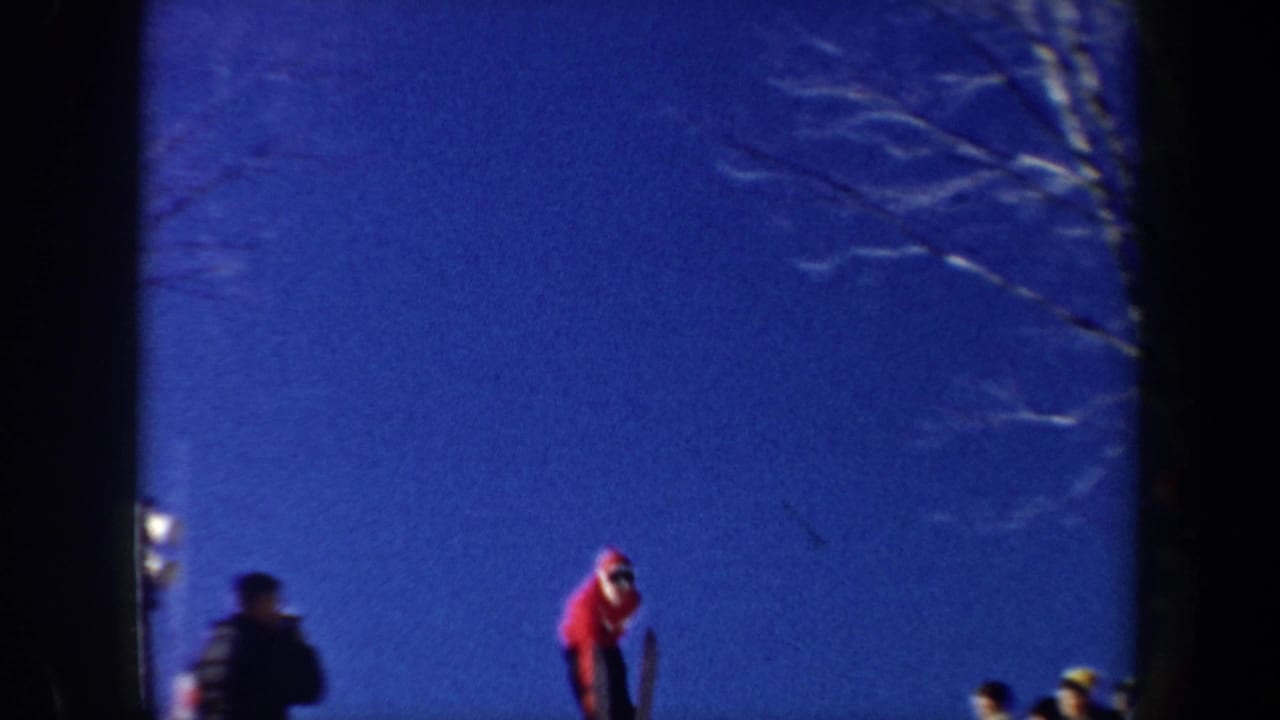 Skier in a red jacket mid-jump against blue sky with spectators near the slope.