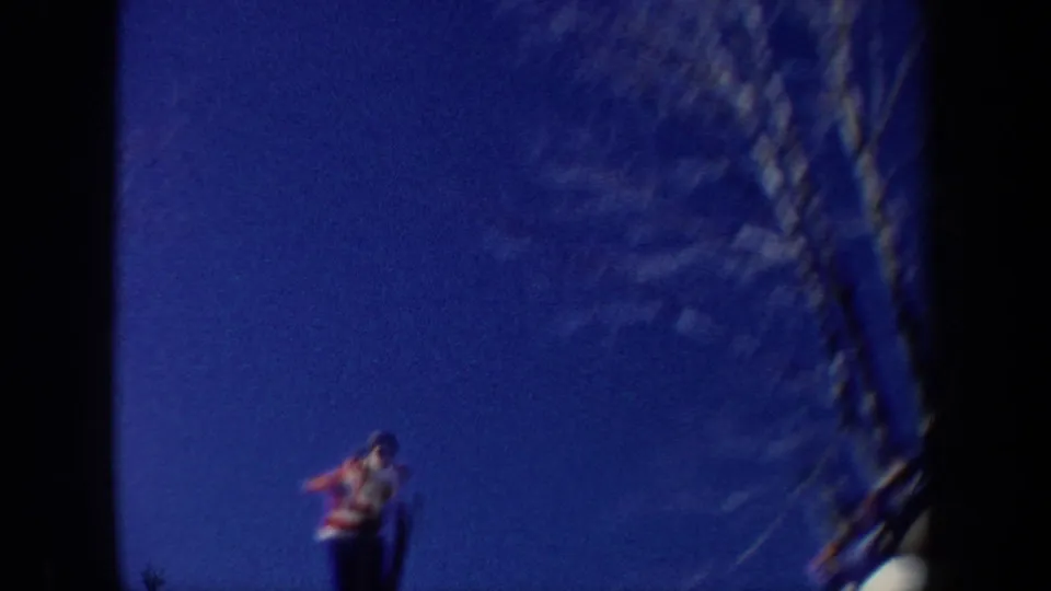 1962 New York home-movie frame with a blurred skier near the lower edge, bare branches, and strong blue sky within visible film borders.
