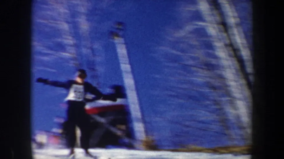 The skier is seen near the landing area with a bib-like front panel, bare trees, snow, and the jump structure behind.