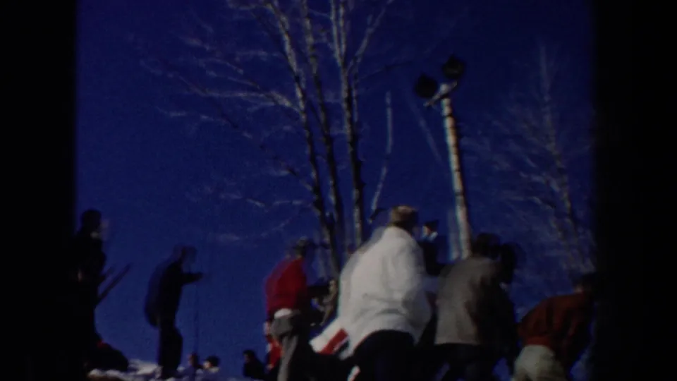 Spectators in winter coats gather near bare trees at the ski site, seen from a handheld 1962 New York home-movie perspective.