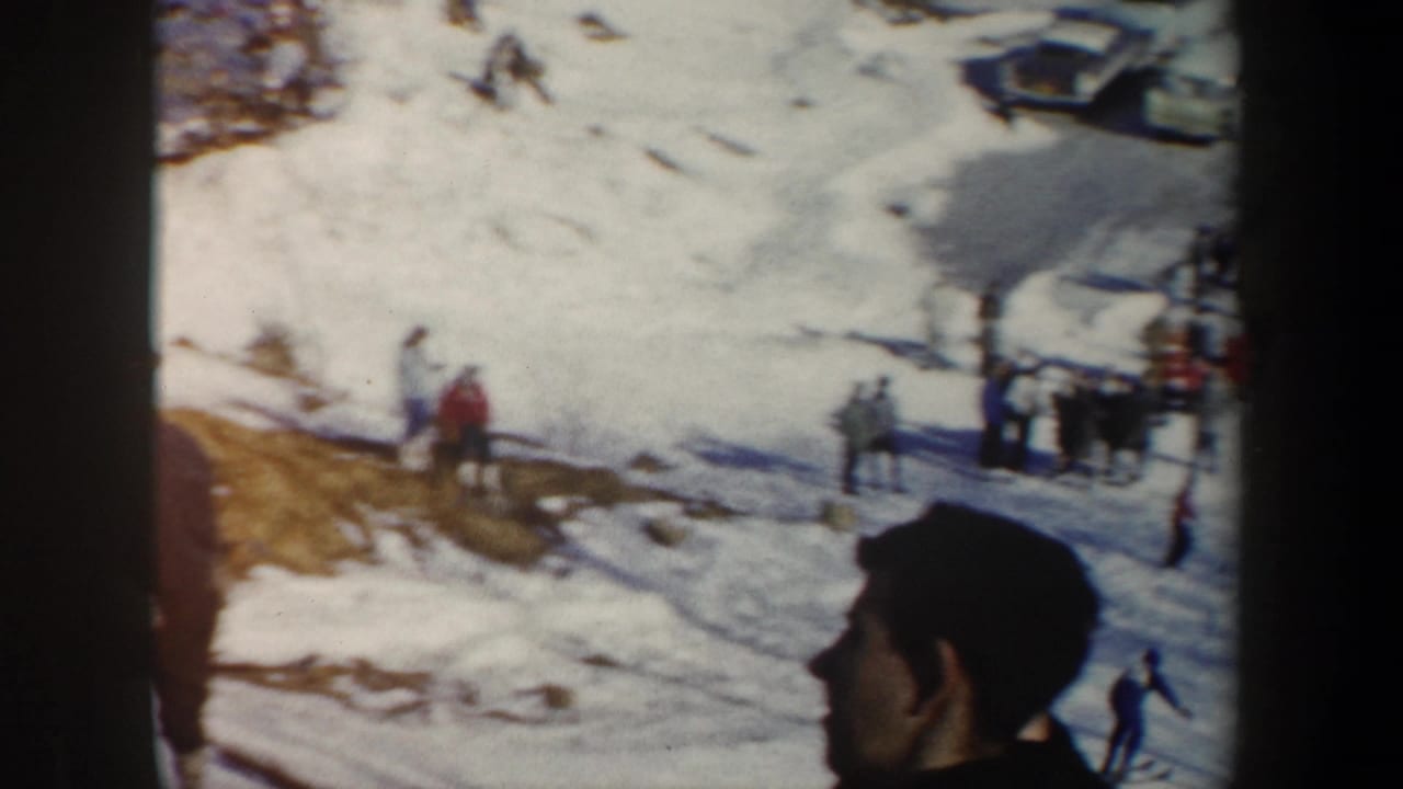1962 New York 8mm frame showing a snowy ski area, distant spectators, parked cars, and a foreground person in profile.