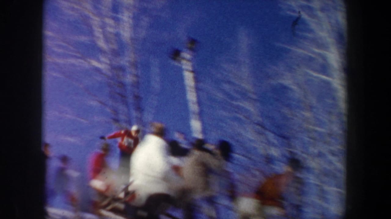 Blurred skier and spectators beneath a blue sky near bare winter trees and light poles.