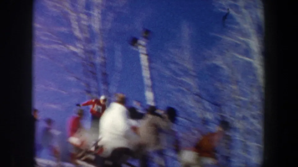 A motion-blurred 1962 New York ski jumping moment with spectators, bare trees, blue sky, and slope-side lighting visible.
