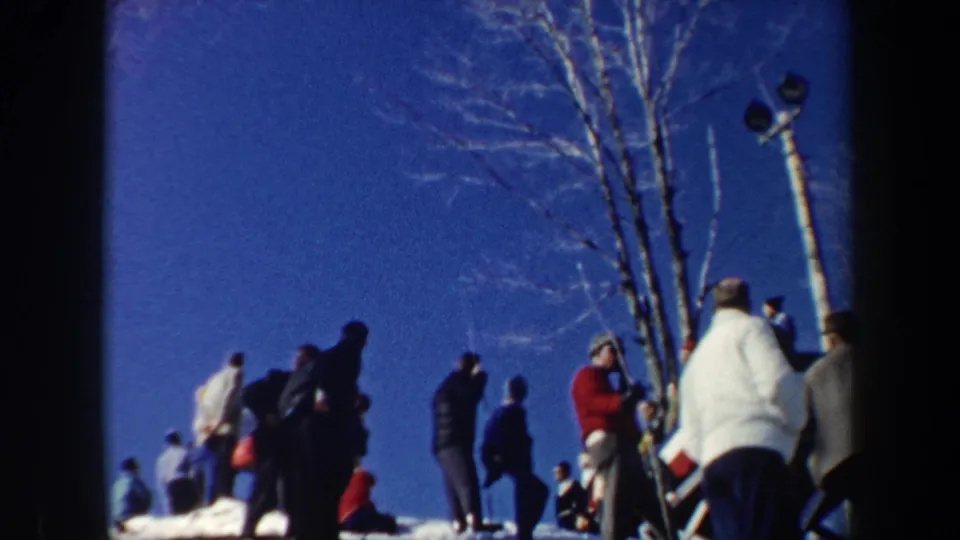 1962 New York home-movie view of bundled spectators standing on a snowy slope near trees and light poles during a ski event.