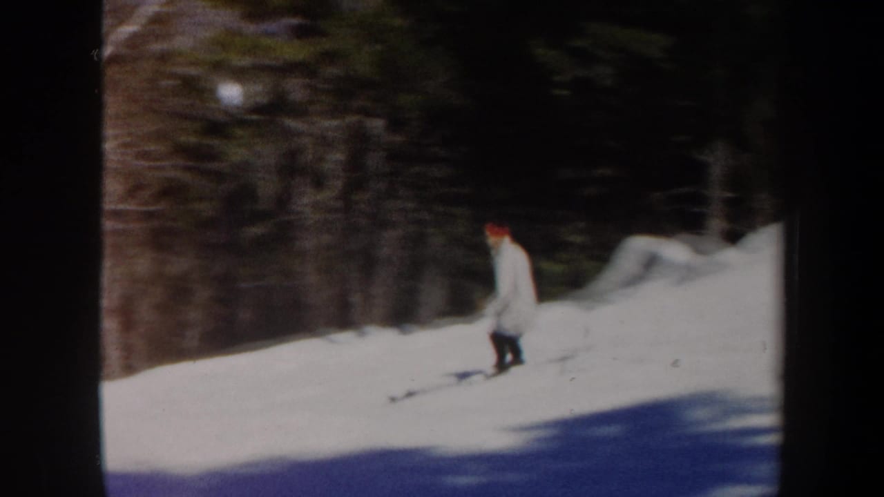 1962 Vermont 8mm frame showing a skier in a light coat and red hat on a gentle snowy slope beside trees.