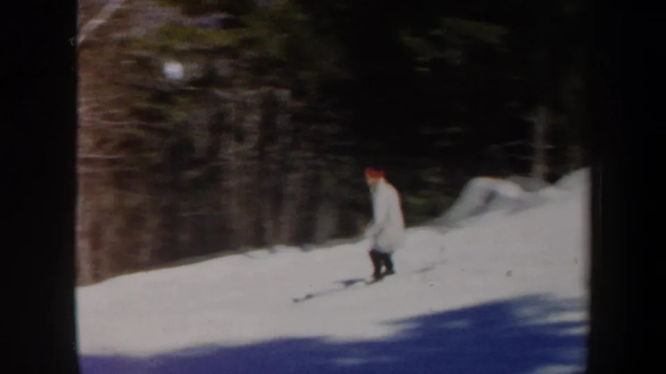 1962 Vermont 8mm frame showing a skier in a light coat and red hat on a gentle snowy slope beside trees.