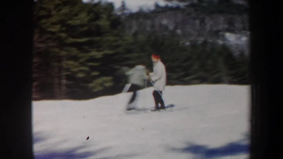 1962 Vermont home-movie frame with two skiers on a gentle snow slope near evergreen woods.
