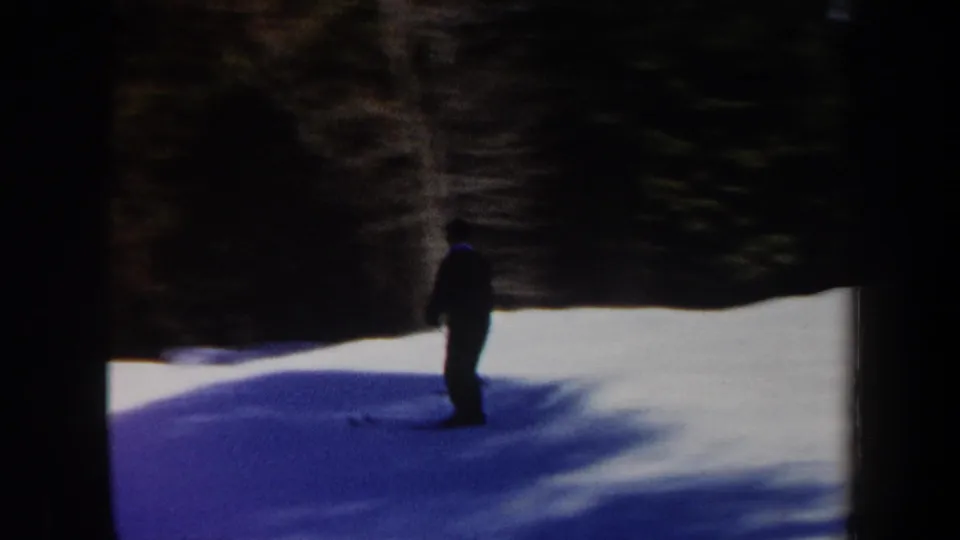 1962 Vermont 8mm frame showing a skier in dark winter clothing descending across a snowfield near dense, shadowed trees.