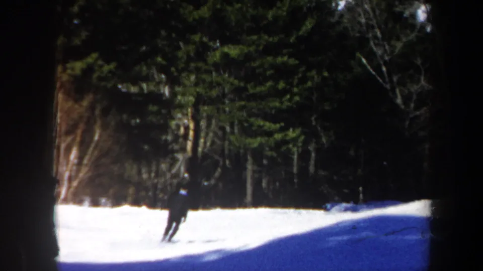 1962 Vermont home-movie frame showing the skier farther down the snowy slope, backed by evergreen woods in bright winter light.