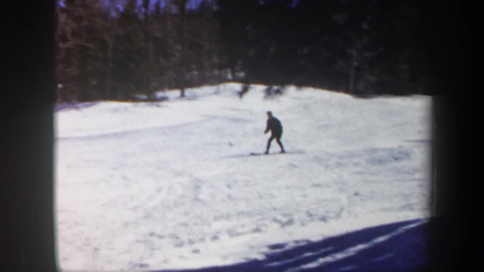 1962 Vermont 8mm frame showing a distant skier moving through an open snowfield below a wooded slope.