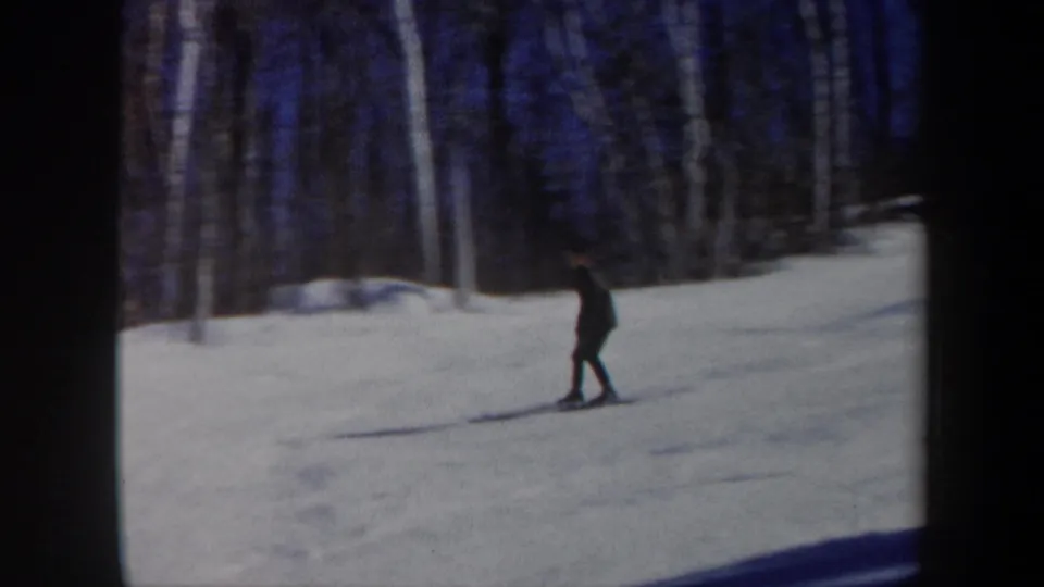 1962 Vermont home-movie frame with a distant skier moving through a quiet snowy forest clearing.