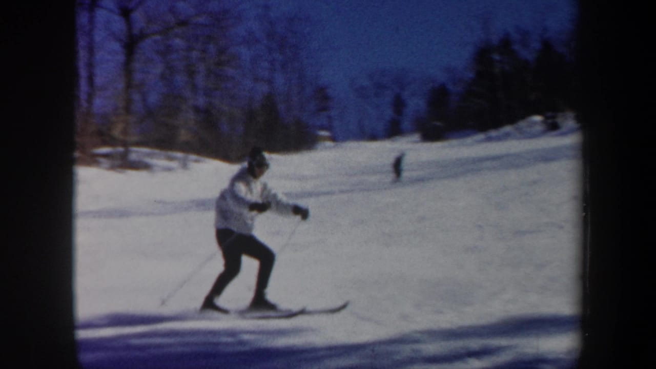 1962 Vermont home-movie frame of a skier crossing a snowy slope, with distant skiers and winter trees behind.